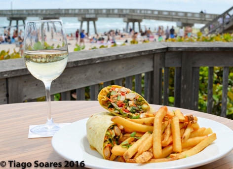 veggie and chicken flour tortilla wrap with a bed of crispy french fries, displayed on a round white plate. Accompanied by a icy glass of white wine. The background is a beach pier on a sunny summer day in Florida. Photographed by Tiago with the Shots.