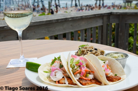 Fish soft tacos with lettuce, lime, guacamole, and pico de gallo. Paired with a side of brown rice displayed on a round white plate. Accompanied by a icy glass of white wine. The background is a beach pier on a sunny summer day in Florida. Photographed by Tiago with the Shots.