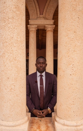 Gentleman in a burgandy suit standing with his hands clasped in front of him. He is standing behind two peach colored pilliars. Photographed by Tiago with the Shots.