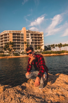Young man posing on beach rocks in Deerfield Beach, Florida. He is squatting down on the rocks in front of a cream colored hotel building across the crystal blue water. Photographed by Tiago with the Shots.