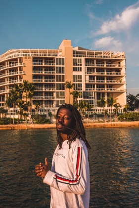 Black man with dreads wearing white and red Tommy Hilfiger long sleeve. Beautiful background with a tan hotel building on a Miami beach shore. Man is looking into the photographers camera with his hands clasped. Photographed by Tiago with the Shots.