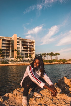 Black man with dreads wearing white and red Tommy Hilfiger long sleeve. Beautiful background with a tan hotel building on a Deerfield beach shore. Man is squatting on a Florida beach rock- looking off into the sunset. Photographed by Tiago with the Shots.