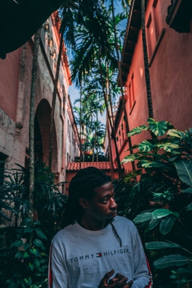Black man with dreads wearing white and red Tommy Hilfiger long sleeve. Beautiful background with a salmon colored building and healthy palm trees and green shrubs. Photographed by Tiago with the Shots.